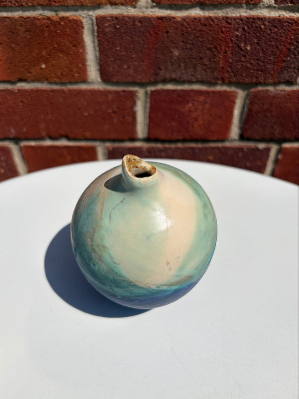 A small round ceramic vase with a glossy glaze in shades of teal, cream, and blue, featuring a small spout and photographed on a white surface with a red brick wall background.